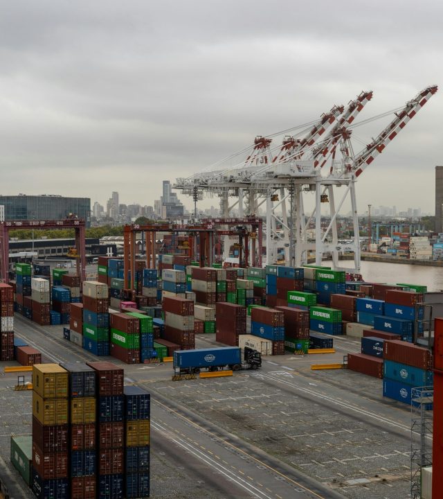 Overhead shot of a bustling container port with cranes and stacked shipping containers under a cloudy sky.