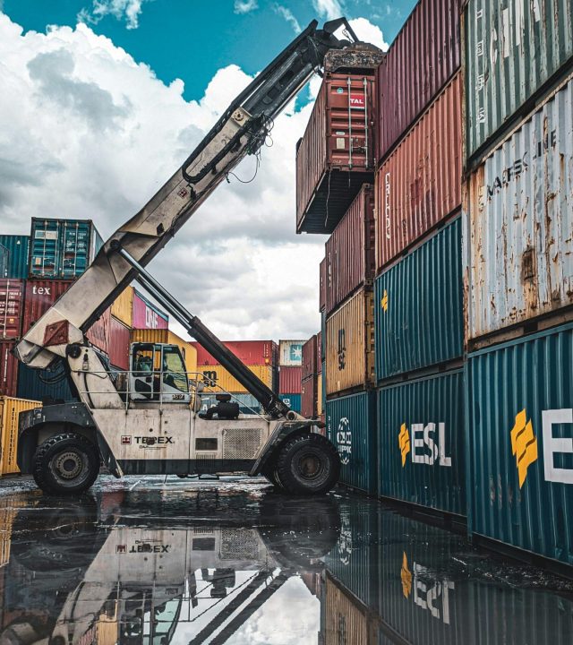 A crane loader stacking colorful shipping containers at a port with reflection in water.